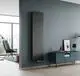 Vertikaler Kermi-Heizkörper in dunklem Design an einer grauen Wand, daneben ein Sideboard mit einem Bild und einer Lampe, Raum mit einem Fenster und einem Sessel.